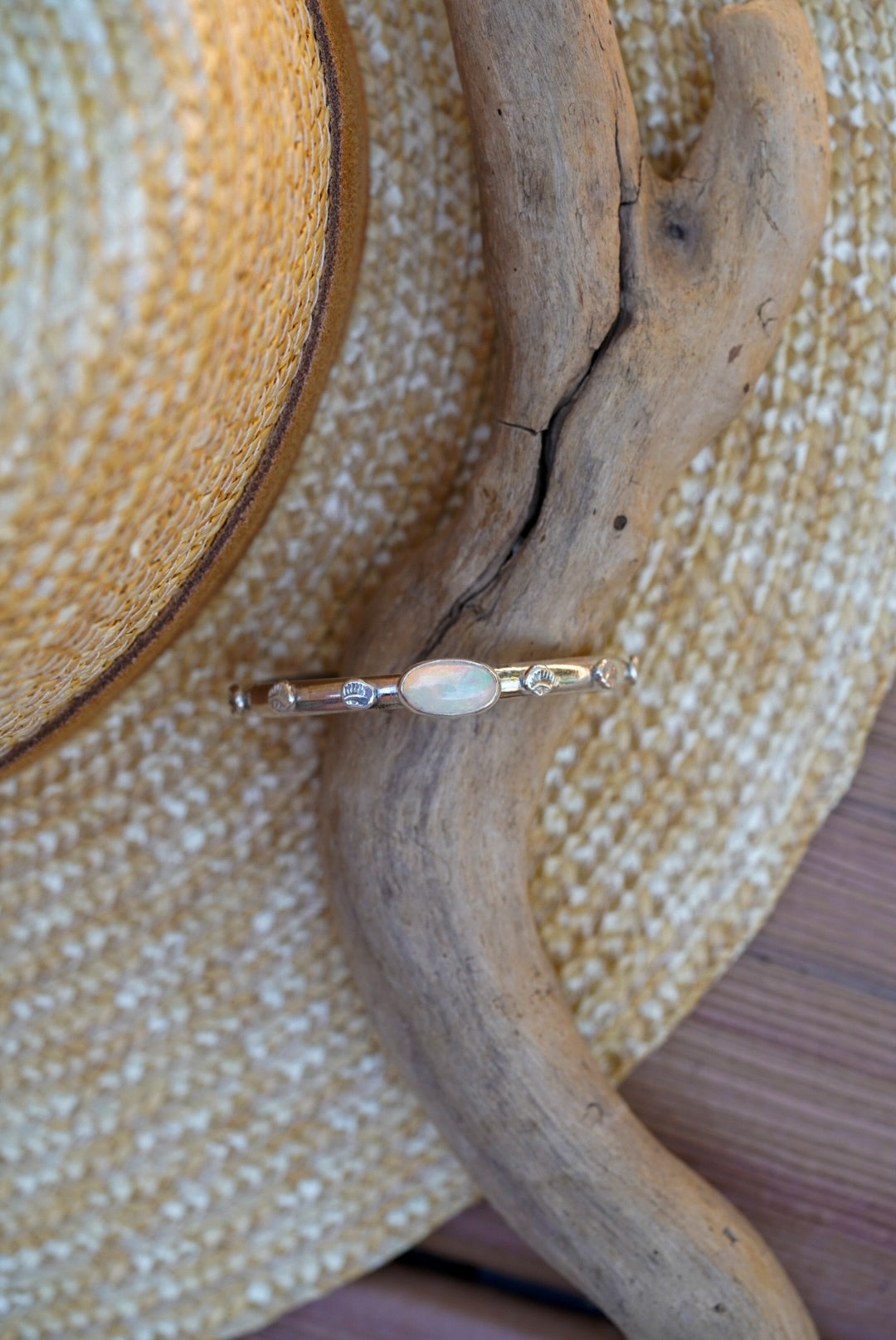 A silver bracelet with a central white opal stone, displayed on a piece of driftwood over a woven straw hat.