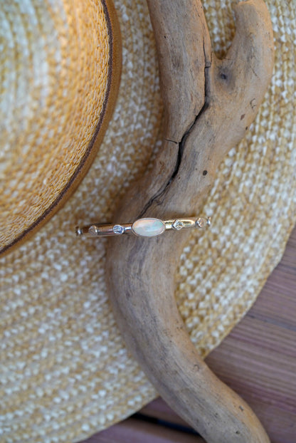 A silver bracelet with a central white opal stone, displayed on a piece of driftwood over a woven straw hat.