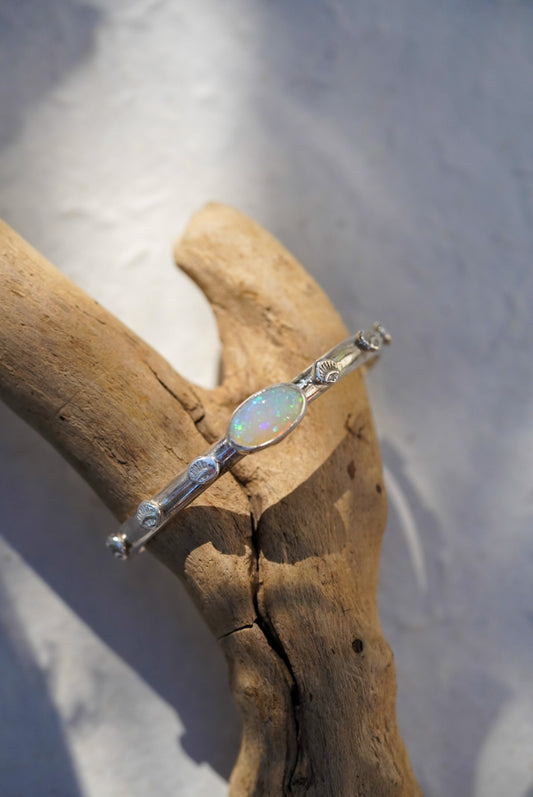 A silver bracelet with a central iridescent opal stone and small stamped details, displayed on a piece of driftwood against a light gray background.