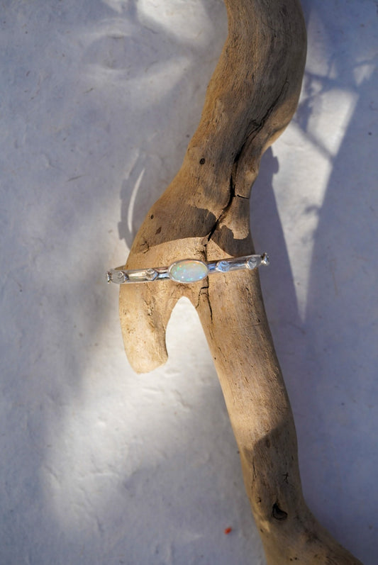 A silver cuff with a central iridescent opal stone, displayed on a piece of driftwood against a white textured background.