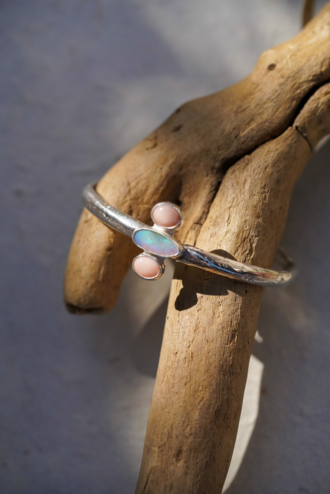 A sterling silver cuff bracelet with a central opal inlay and small pink cabochon stones, displayed on a piece of driftwood.