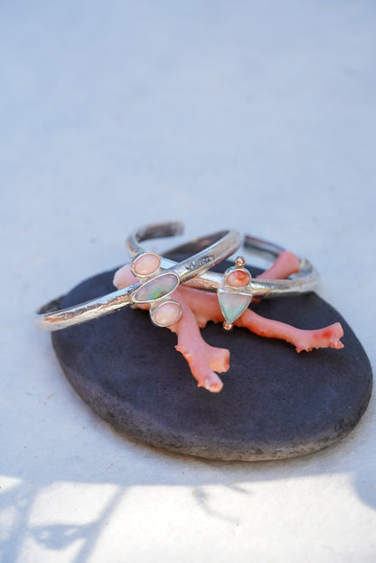two handmade silver cuffs with coral and opal accents displayed on a dark stone against a light background.