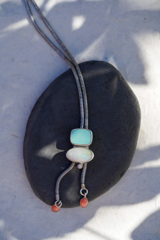 A silver bolo-style necklace with a light blue stone, a opal stone, and small coral-red beads, displayed on a dark stone against a white background with soft shadows.