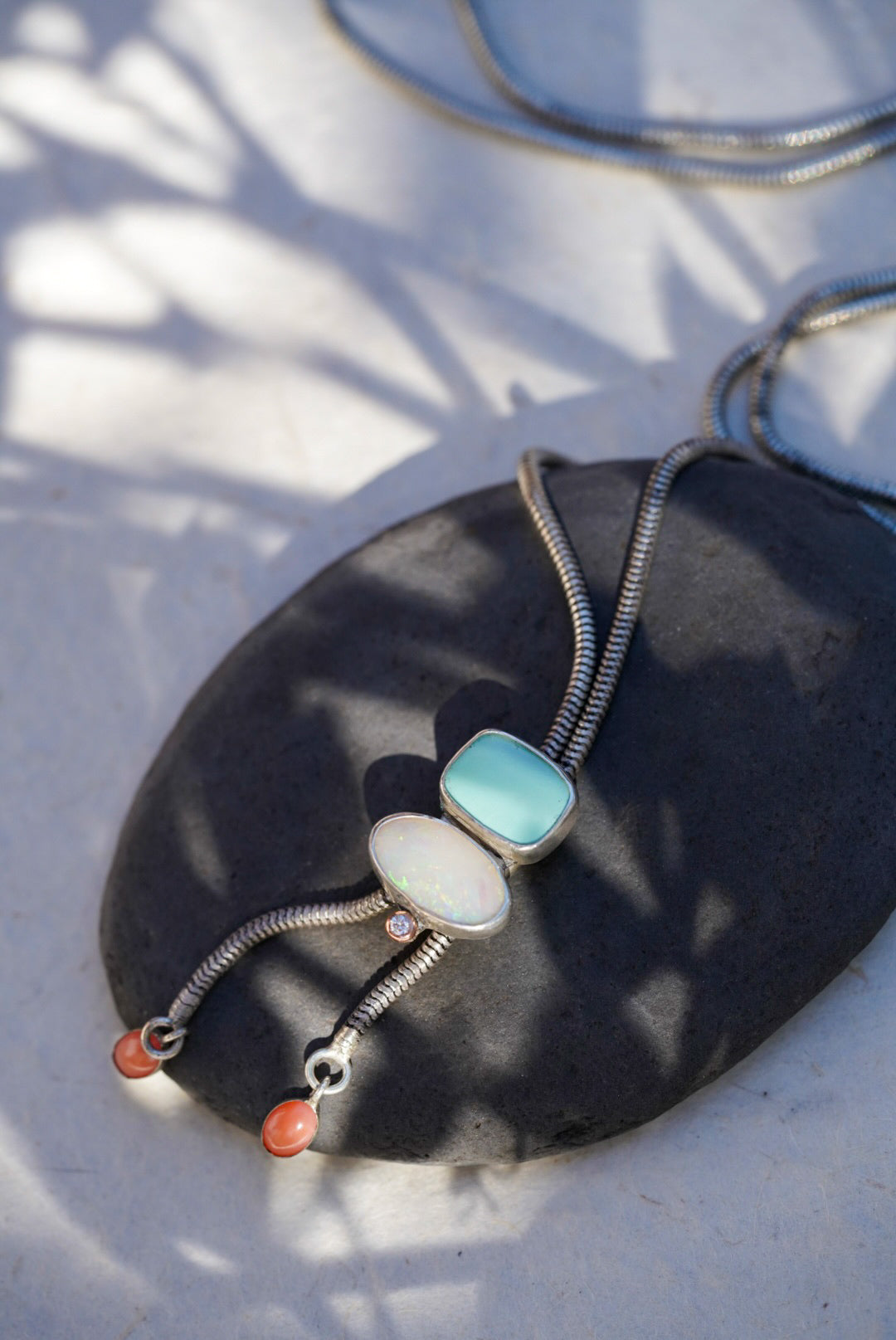 A silver snake chain bolo necklace with a light blue stone, an opal stone, and small coral beads, displayed on a dark stone slab with dappled sunlight.