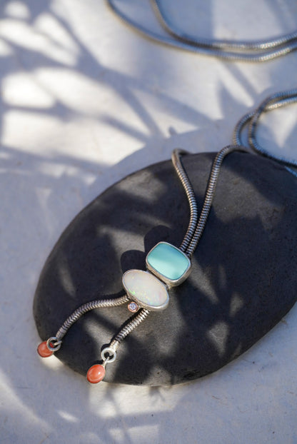A silver snake chain bolo necklace with a light blue stone, an opal stone, and small coral beads, displayed on a dark stone slab with dappled sunlight.