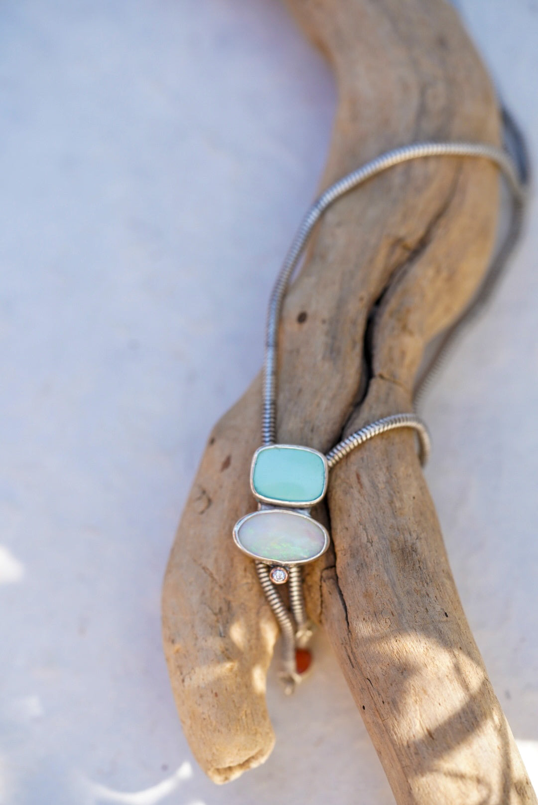 A silver bolo with a rectangular light blue stone and an oval opal stone, displayed on a piece of driftwood against a pale background.