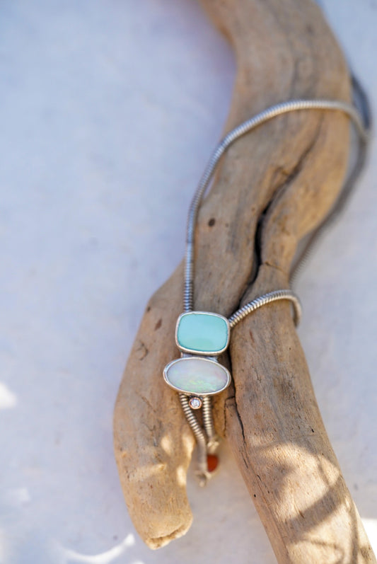 A silver bolo with a rectangular light blue stone and an oval opal stone, displayed on a piece of driftwood against a pale background.