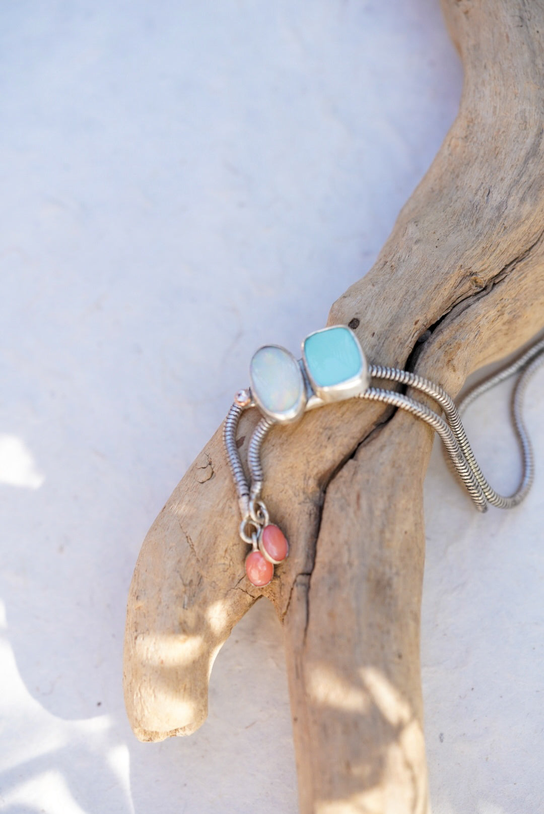 A silver snake chain bolo with a oval opal stone, a square light blue stone, and two small pink beads, displayed on a piece of driftwood against a white background.