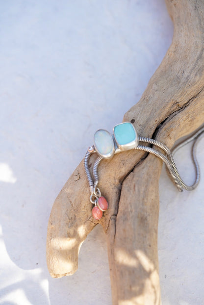 A silver snake chain bolo with a oval opal stone, a square light blue stone, and two small pink beads, displayed on a piece of driftwood against a white background.
