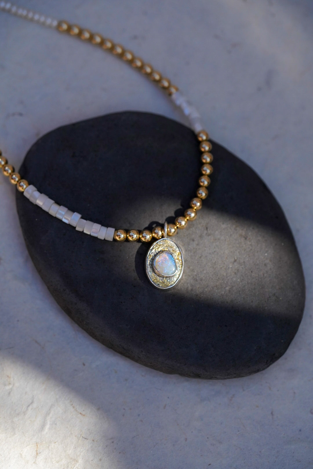 A beaded necklace with gold and pearl beads, featuring a central oval pendant with an opal stone, displayed on a dark stone prop against a light textured background.