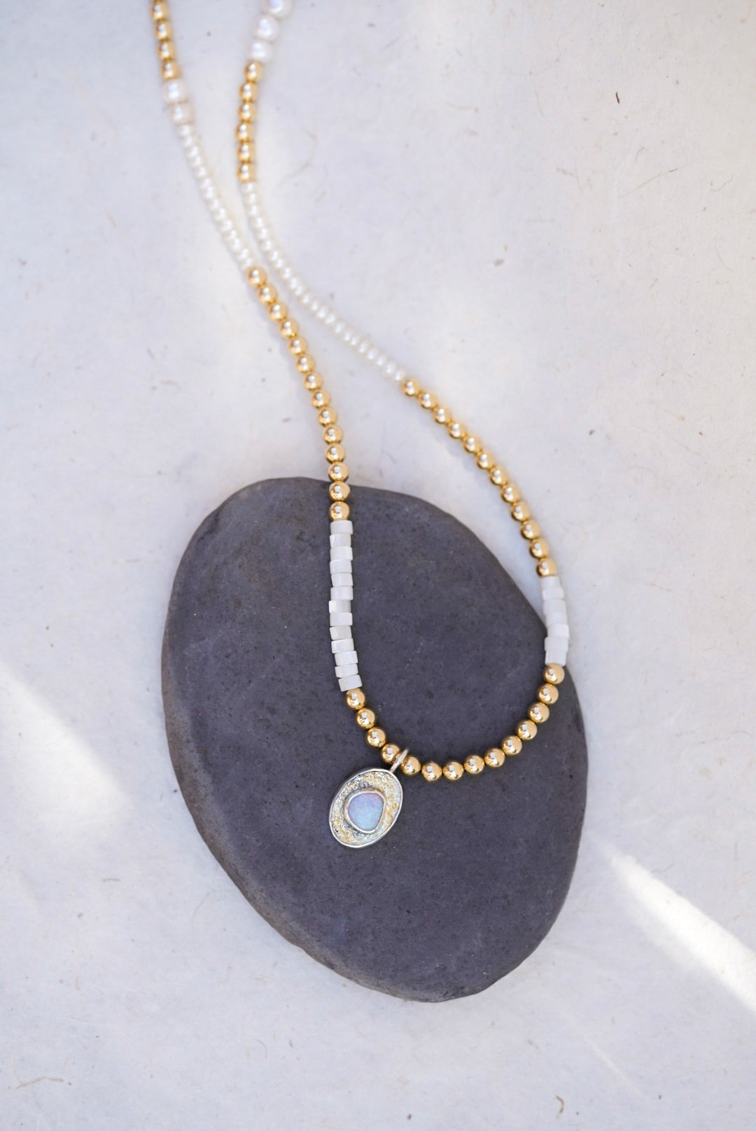 A delicate beaded necklace with gold and pearl beads, featuring a small oval pendant with a opal stone, displayed on a dark gray stone against a light background.