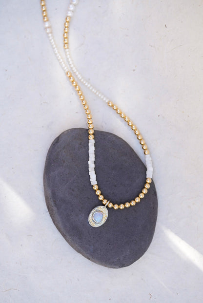 A delicate beaded necklace with gold and pearl beads, featuring a small oval pendant with a opal stone, displayed on a dark gray stone against a light background.