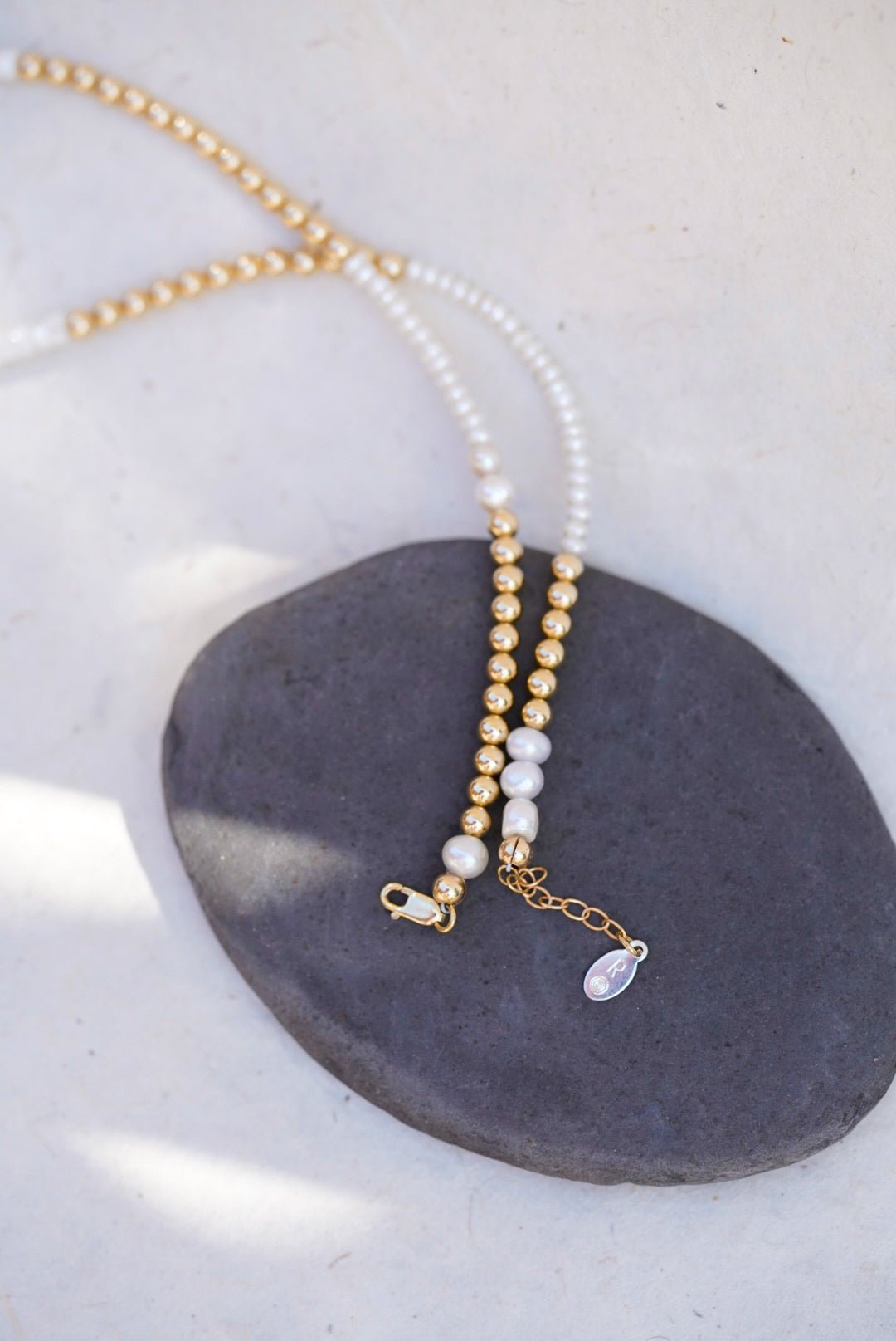 A delicate beaded necklace with pearl beads and gold spacer beads, displayed on a dark gray stone against a light beige background, showing the lobster clasp.