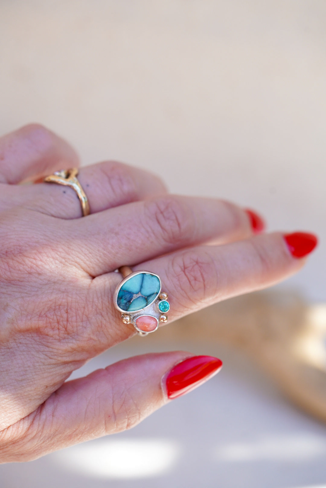 A close-up of a hand wearing a gold ring with a large marbled turquoise cabochon and smaller pink and green stones, against a soft beige background.