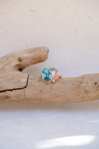 A handmade ring with a large turquoise-blue stone, a coral-pink stone, and a small blue stone, displayed on a piece of driftwood against a light beige background.