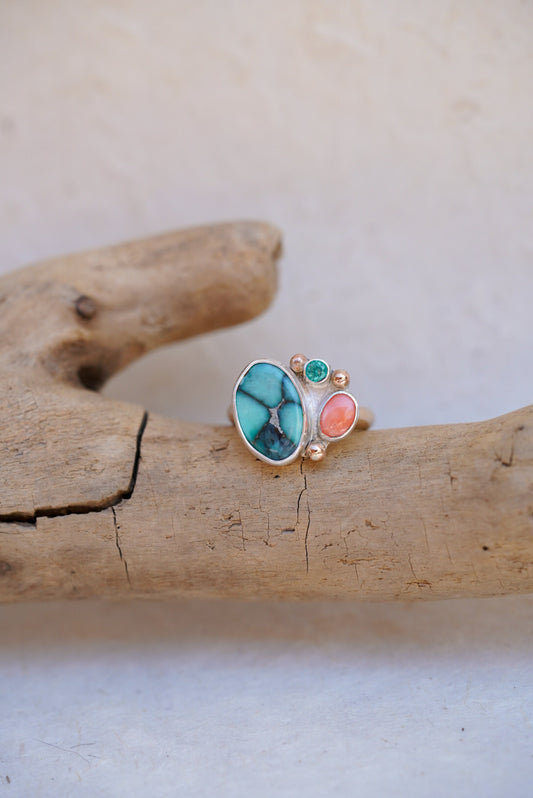 A handmade silver ring with a turquoise stone and coral stone, displayed on a piece of driftwood against a light beige background.