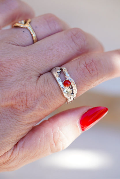 A close-up of a hand wearing a textured silver ring with a central red coral gemstone, alongside other gold rings, against a soft beige background.