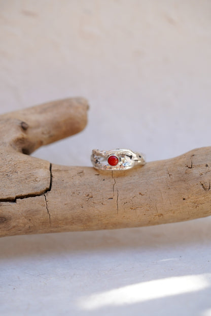 A silver ring with a small red coral stone, displayed on a piece of natural driftwood against a light beige background.