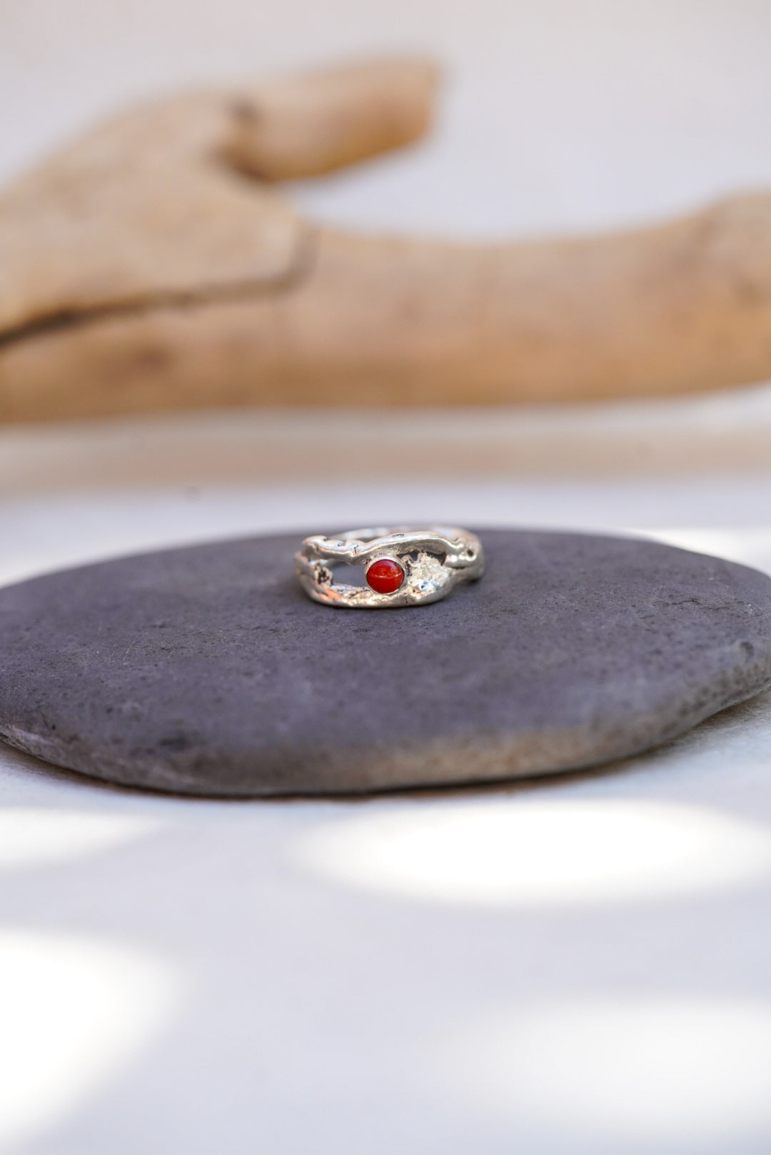 A silver ring with a small round red coral gemstone, displayed on a dark gray stone with a blurred wooden prop in the background.