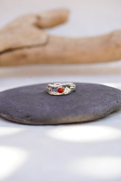 A silver ring with a small round red coral gemstone, displayed on a dark gray stone with a blurred wooden prop in the background.