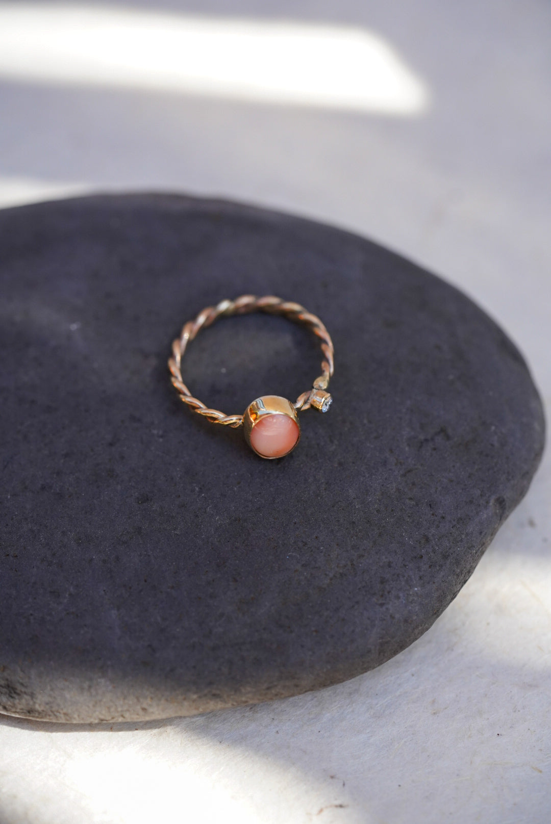 A twisted gold ring with an oval pink coral stone and a small diamond stone, displayed on a dark gray stone surface.