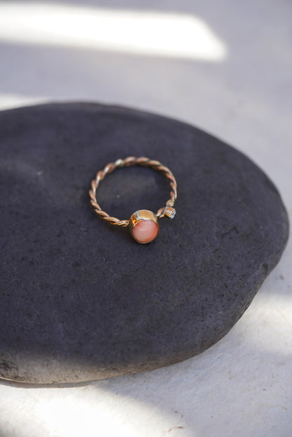 A twisted gold ring with an oval pink coral stone and a small diamond stone, displayed on a dark gray stone surface.