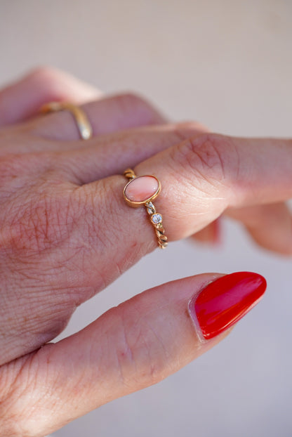 A close-up of a hand wearing a delicate gold ring with a small oval pink coral stone and diamond accent stone, alongside other rings and a red-painted fingernail.