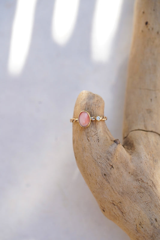 A delicate gold ring with a pink oval coral stone and a small diamond accent, displayed on a piece of driftwood against a bright, softly lit background.