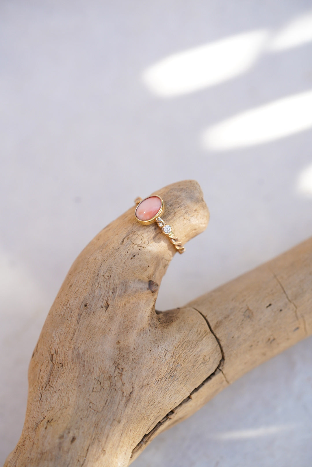 A gold ring with a pink oval coral stone and small diamond, displayed on a piece of driftwood against a light background.