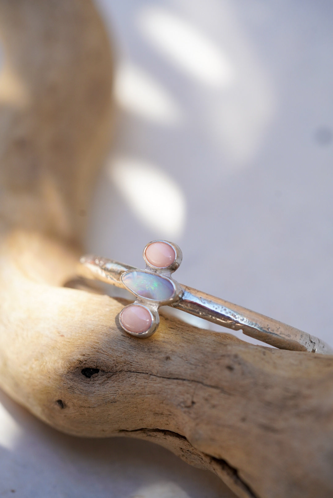A silver cuff with a central iridescent opal and two small pink coral stones, displayed on a piece of driftwood.