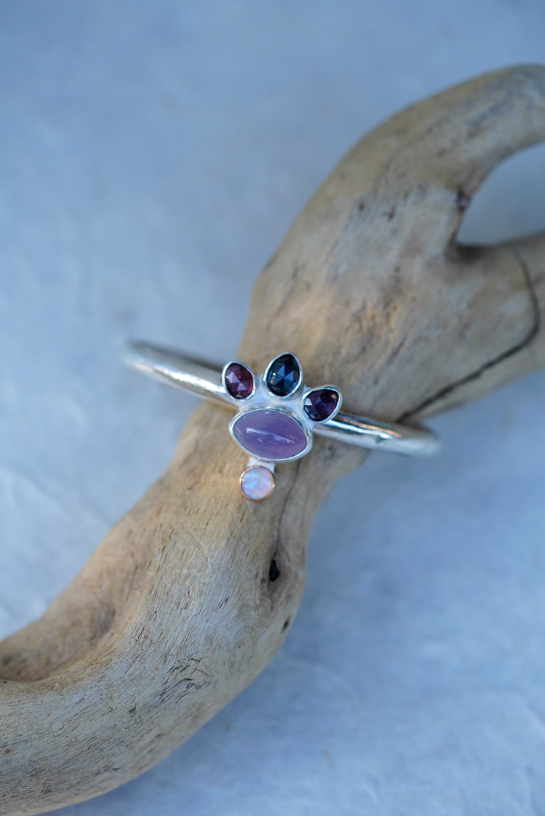A silver cuff with oval purple chalcedony, three faceted spinels, and a small opal, displayed on a piece of driftwood against a light blue background.