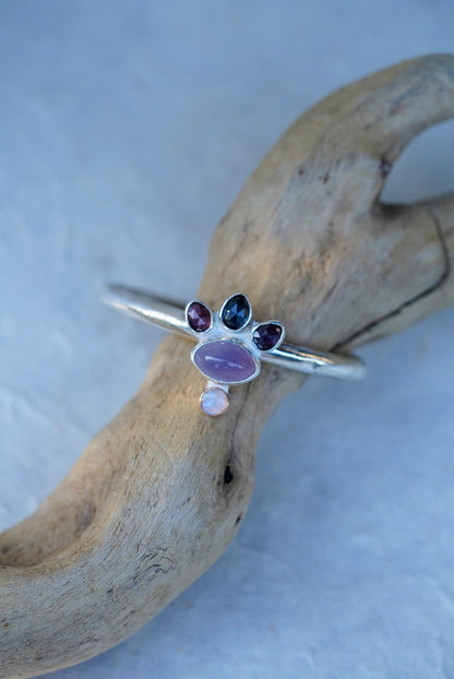 A silver cuff with oval purple chalcedony, three faceted spinels, and a small opal, displayed on a piece of driftwood against a light blue background.