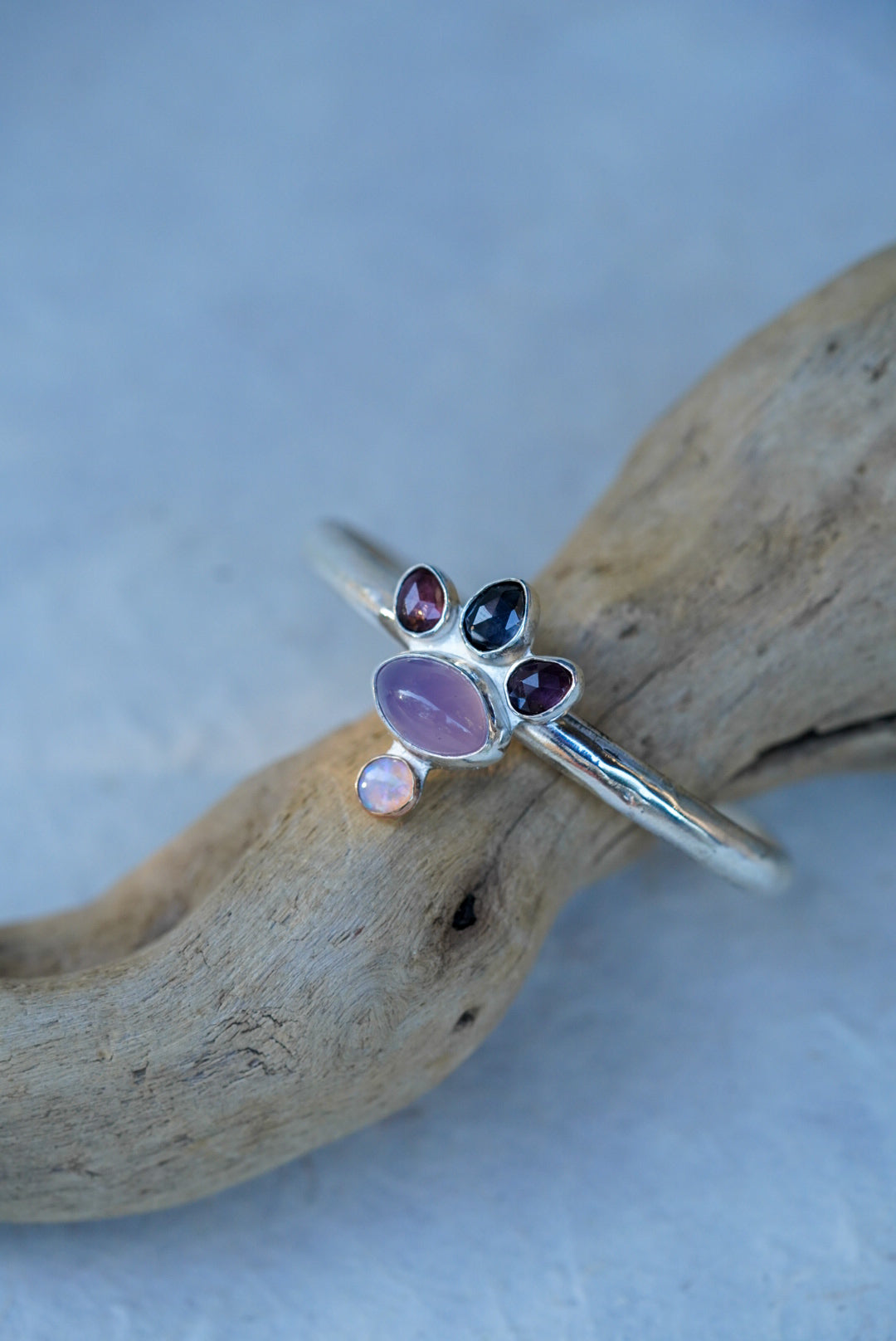 A silver cuff with oval purple chalcedony, three faceted spinels, and a small opal, displayed on a piece of driftwood against a light blue background.