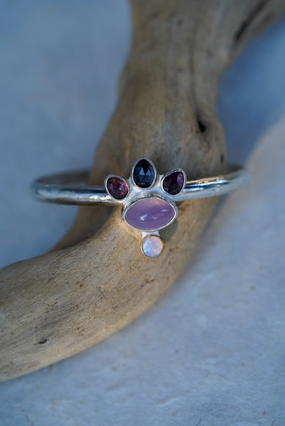 A silver cuff with oval purple chalcedony, three faceted spinels, and a small opal, displayed on a piece of driftwood against a light blue background.