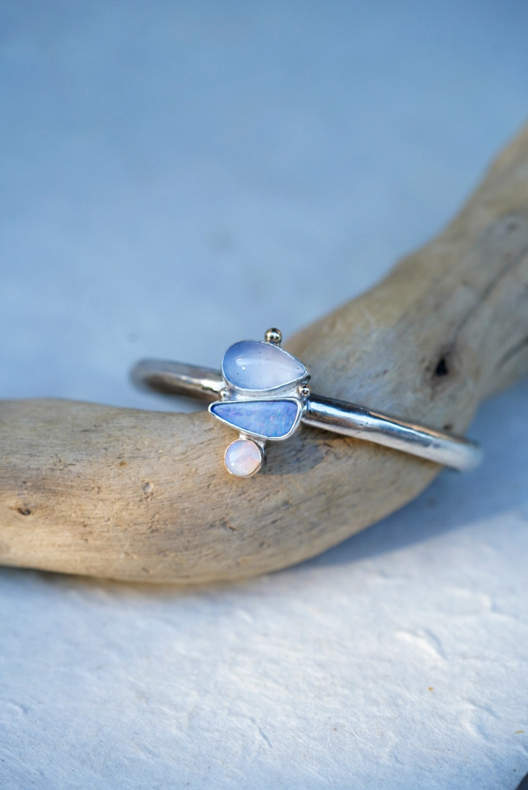 A silver cuff with three stones in shades of light blue and purple, chalcedony and opals, displayed on a piece of driftwood against a light blue background.