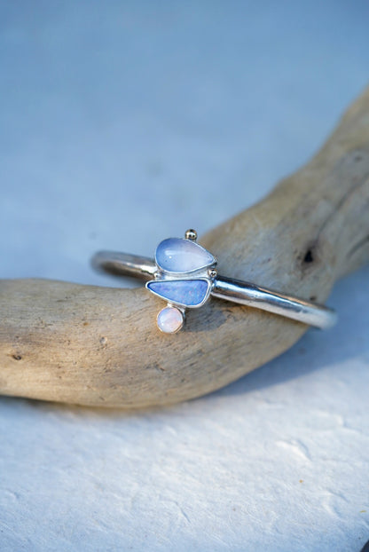A silver cuff with three stones in shades of light blue and purple, chalcedony and opals, displayed on a piece of driftwood against a light blue background.