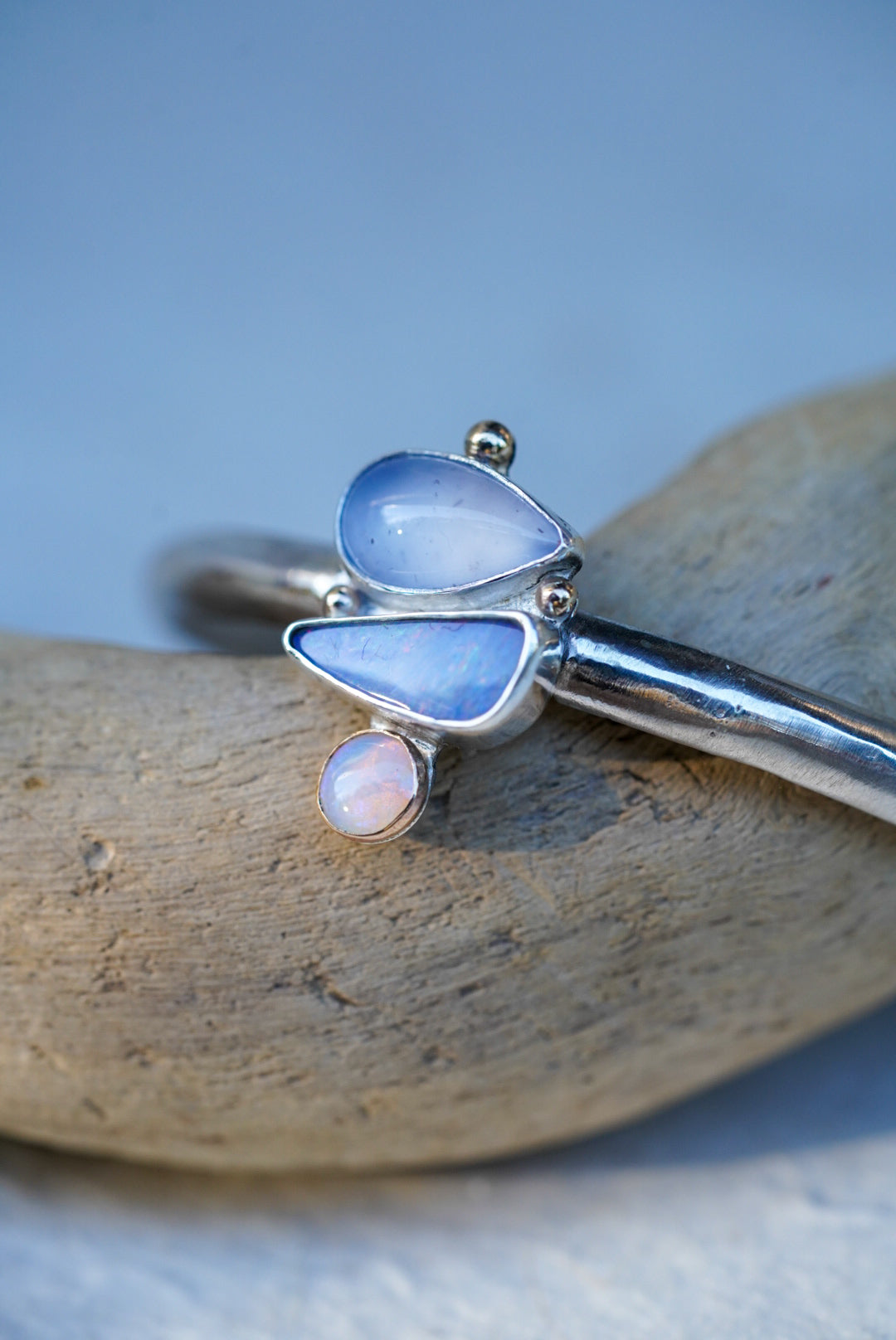 A silver cuff with three stones in shades of light blue and purple, chalcedony and opals, displayed on a piece of driftwood against a light blue background.