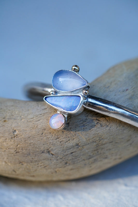 A silver cuff with three stones in shades of light blue and purple, chalcedony and opals, displayed on a piece of driftwood against a light blue background.