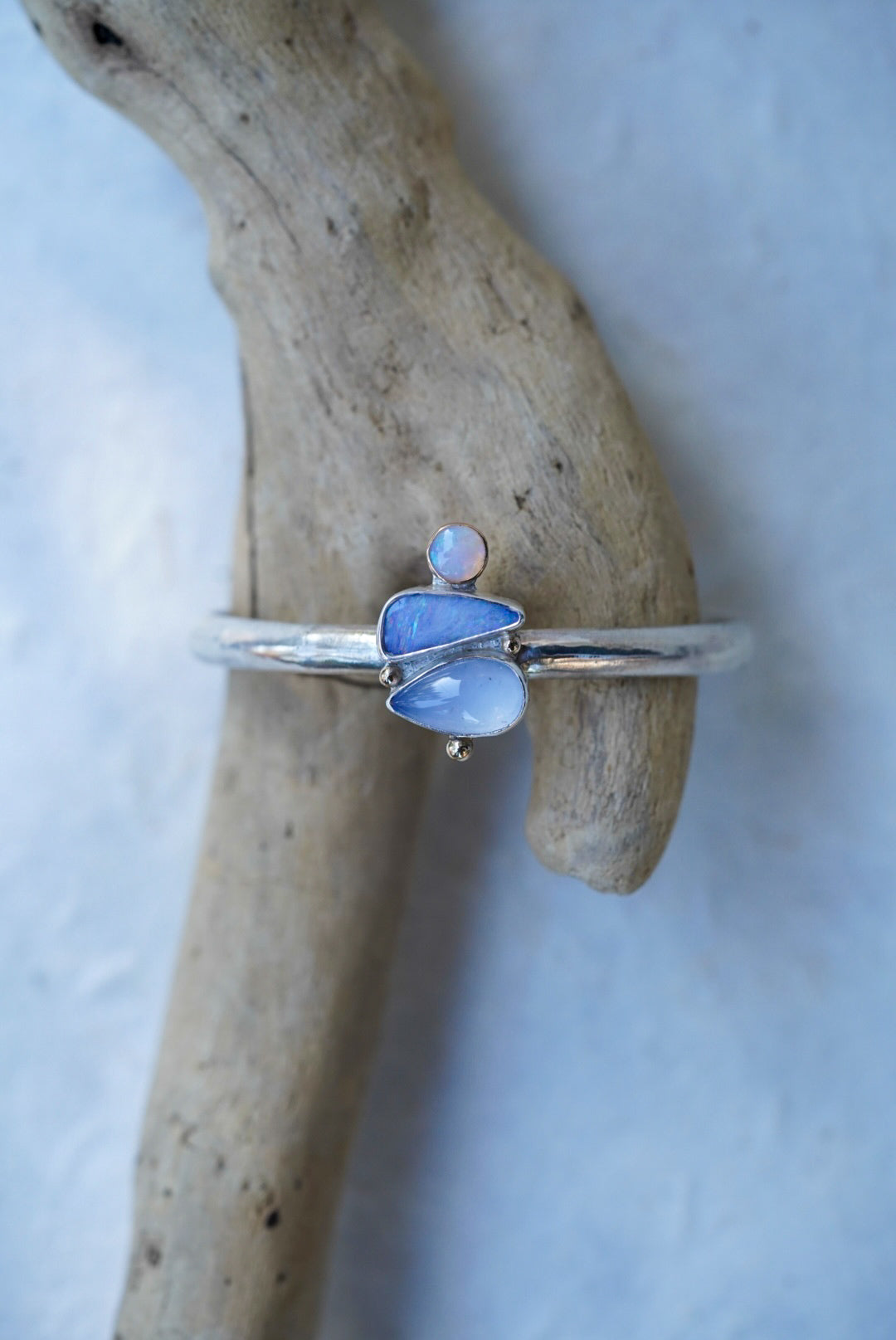 A silver cuff with three stones in shades of light blue and purple, chalcedony and opals, displayed on a piece of driftwood against a light blue background.