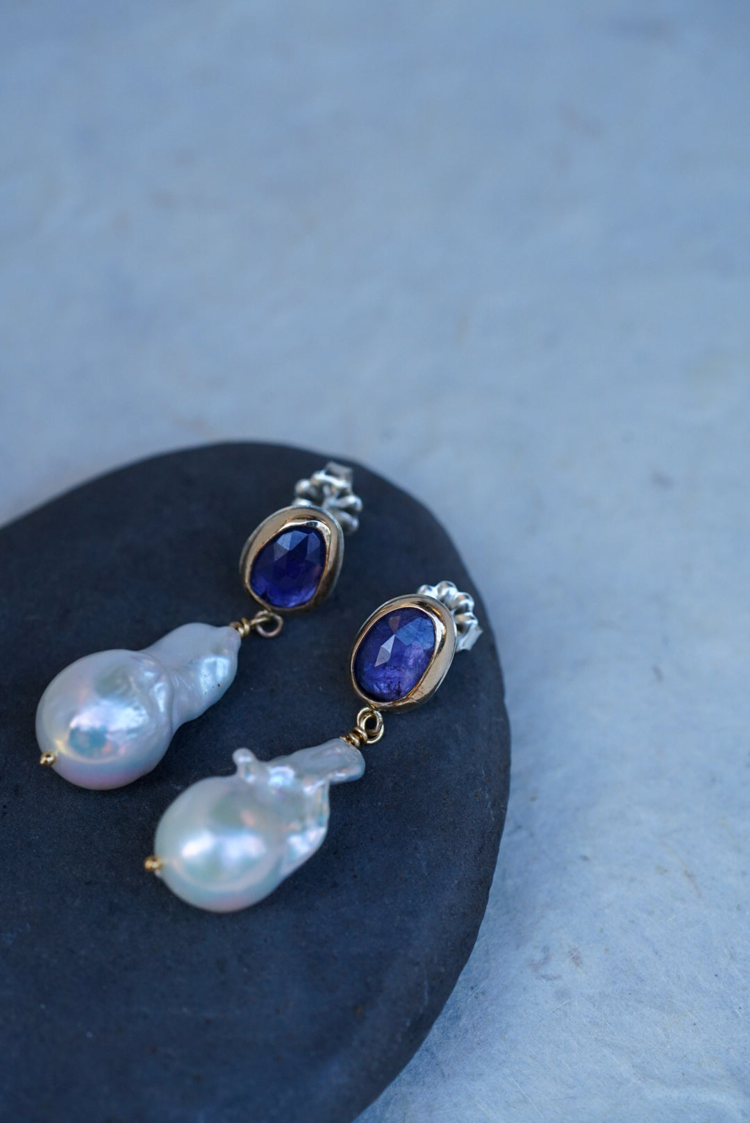 A pair of gold drop earrings with oval tanzanite gemstones at the top and irregular baroque pearl drops below, displayed on a dark stone against a light textured background.