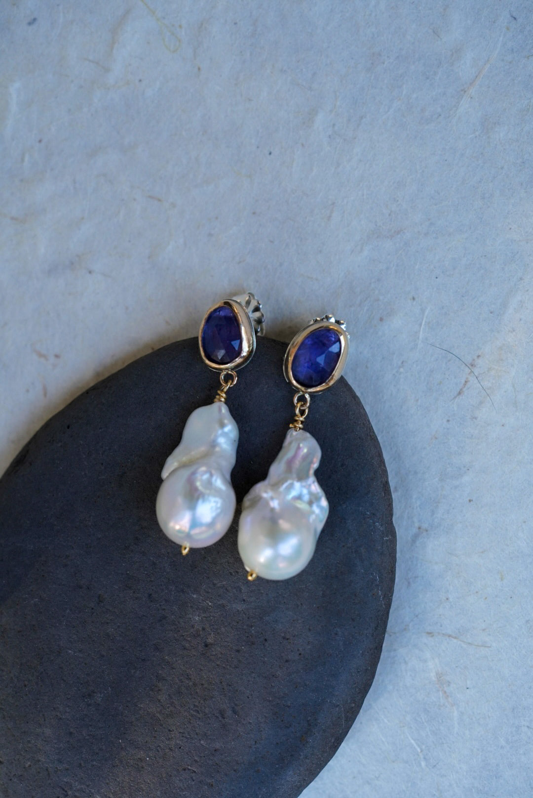 A pair of gold drop earrings with oval tanzanite gemstones at the top and irregular baroque pearl drops below, displayed on a dark stone against a light textured background.