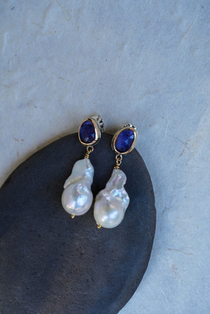 A pair of gold drop earrings with oval tanzanite gemstones at the top and irregular baroque pearl drops below, displayed on a dark stone against a light textured background.