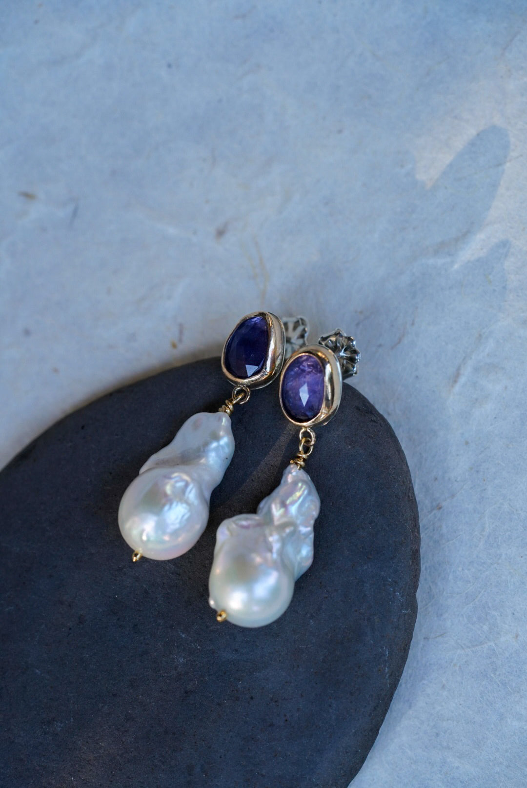 A pair of gold drop earrings with oval tanzanite gemstones at the top and irregular baroque pearl drops below, displayed on a dark stone against a light textured background.