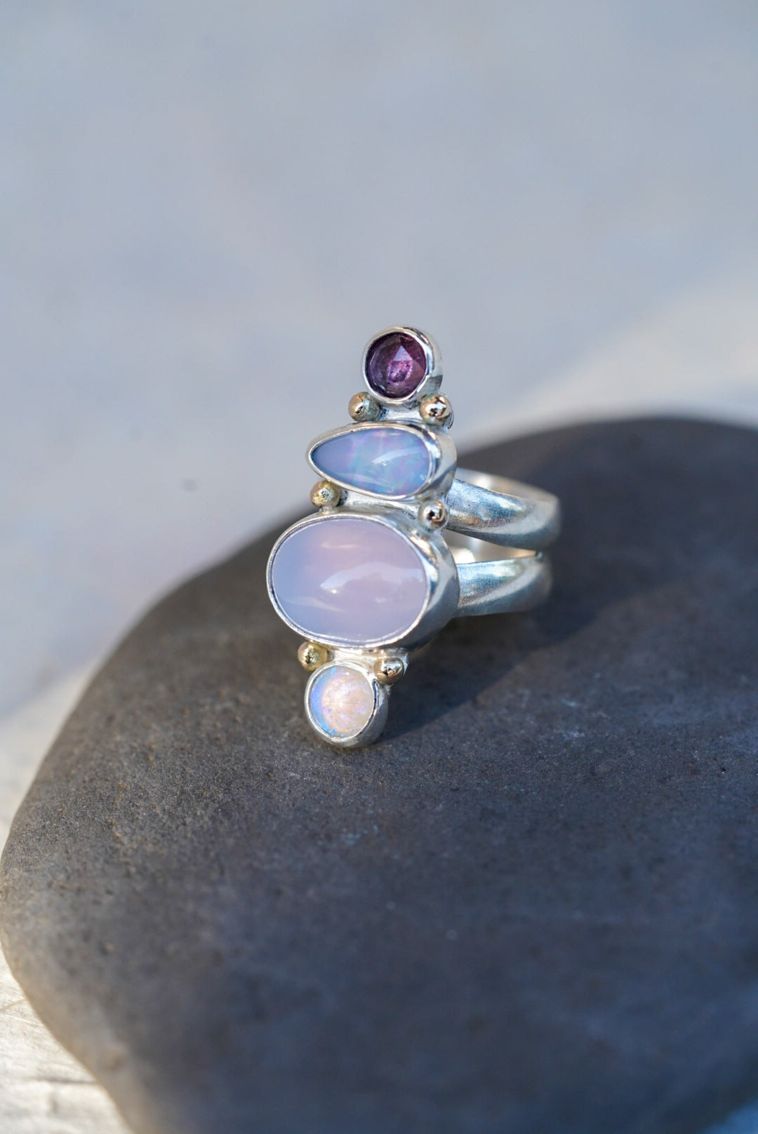 A silver ring featuring a vertical stack of gemstones including a purple chalcedony, opals, and a spinel, displayed on a dark stone surface.