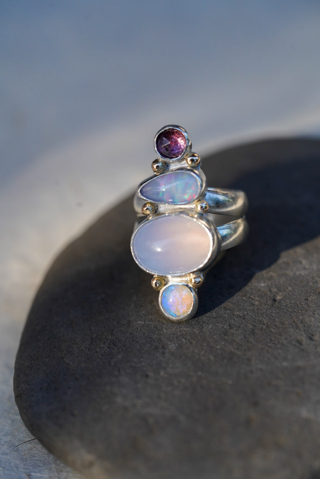 A silver ring featuring a vertical stack of gemstones including a purple chalcedony, opals, and a spinel, resting on a dark stone surface.