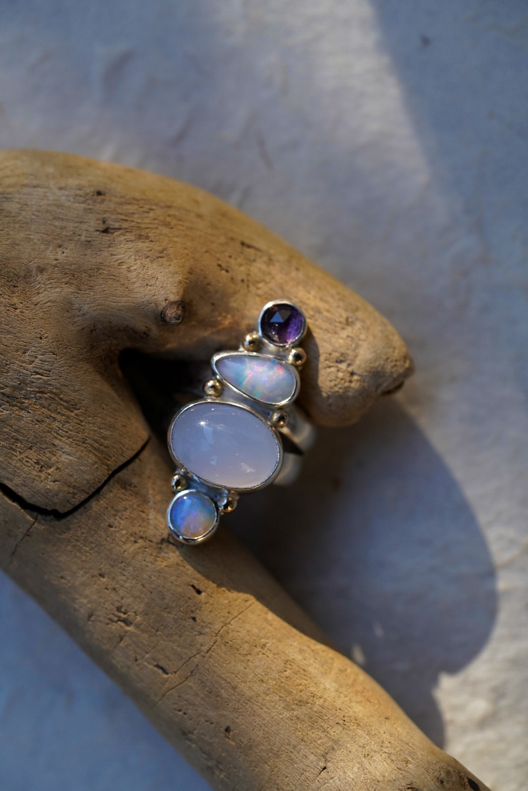 A silver ring featuring a vertical stack of gemstones including a purple chalcedony, opals, and a spinel, displayed on a piece of driftwood.