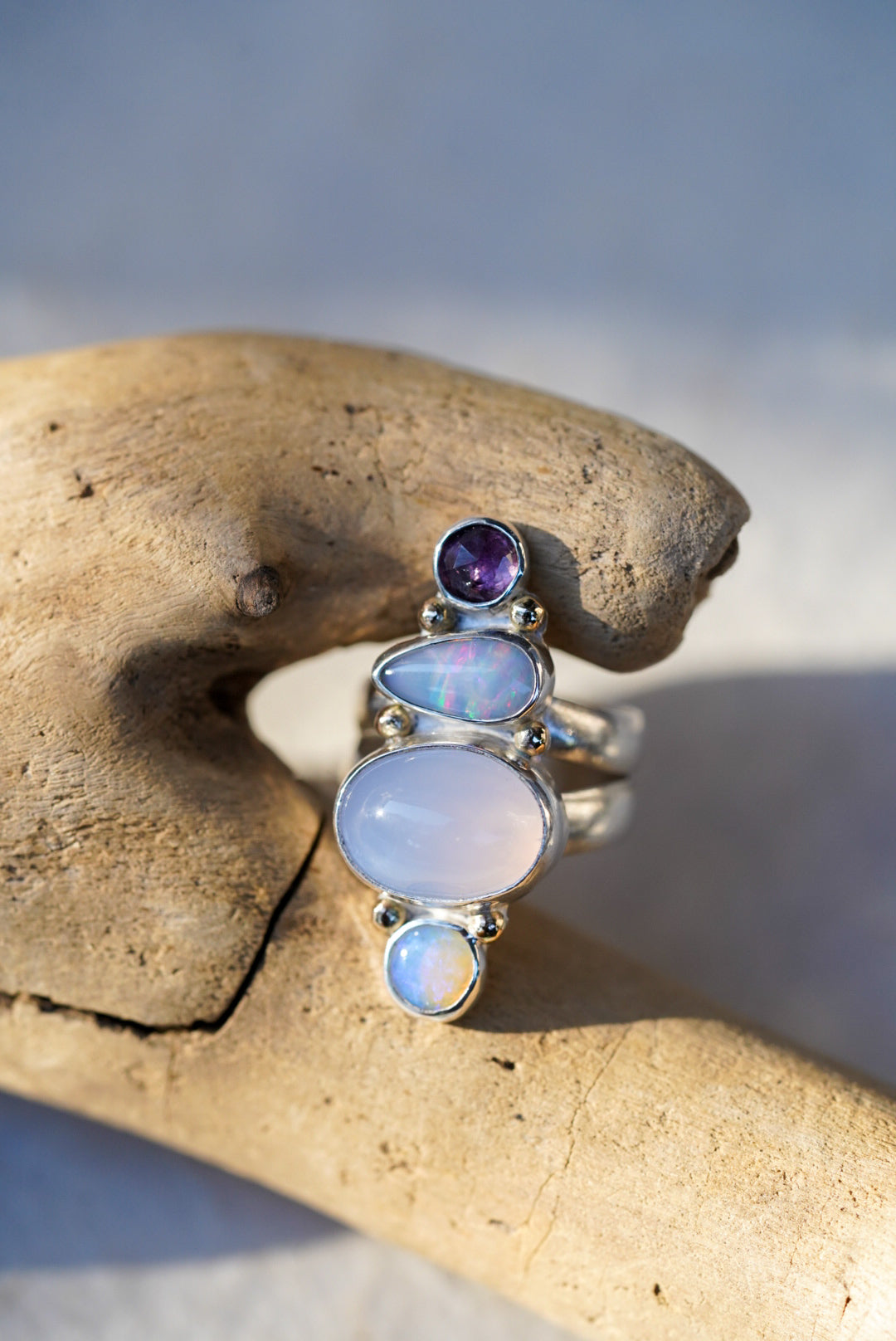 A silver ring featuring a vertical stack of gemstones including a purple chalcedony, opals, and a spinel, displayed on a piece of driftwood.