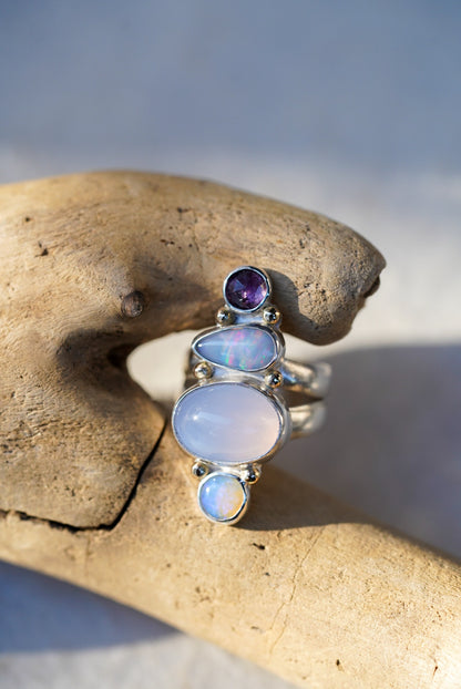 A silver ring featuring a vertical stack of gemstones including a purple chalcedony, opals, and a spinel, displayed on a piece of driftwood.