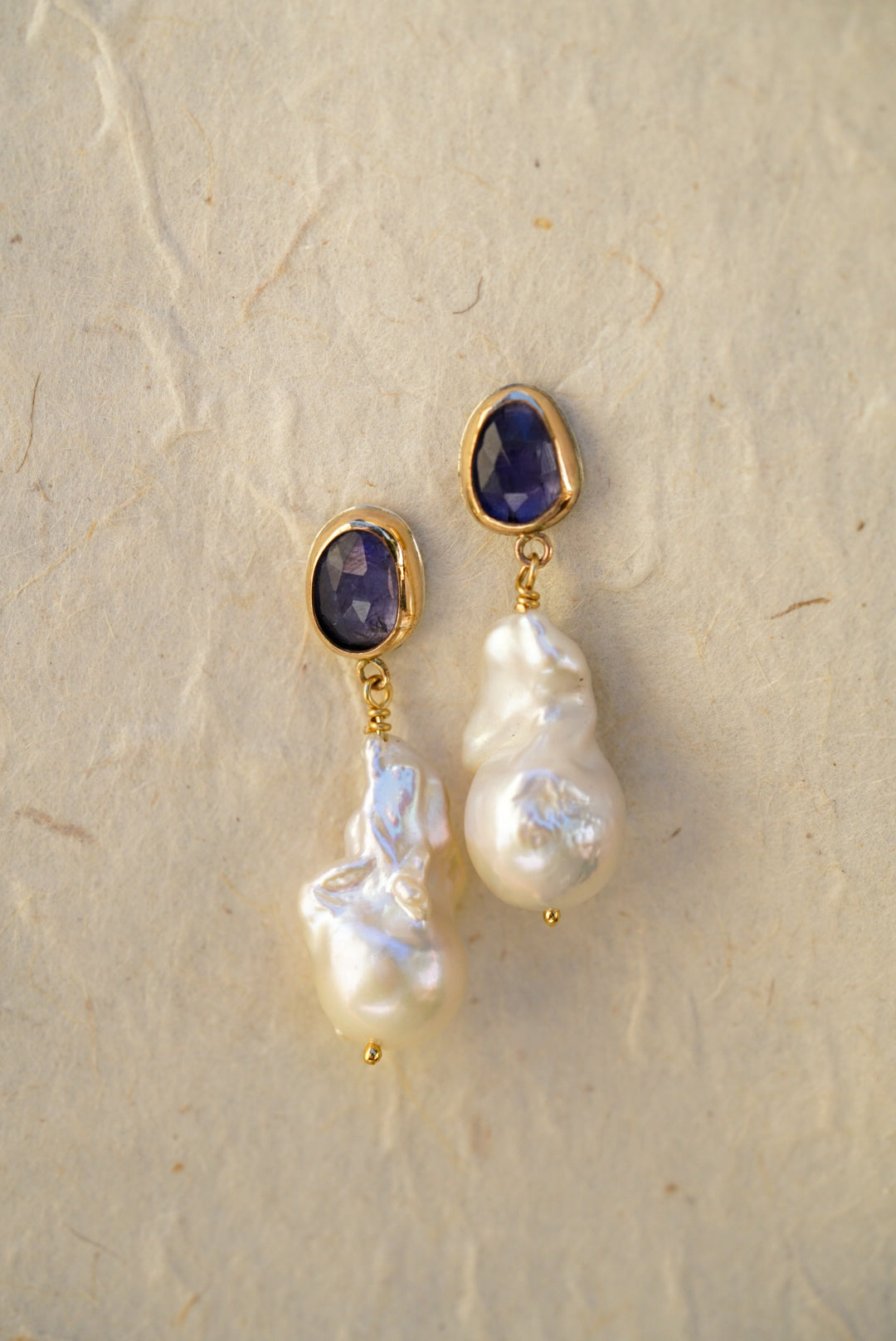 A pair of gold drop earrings with oval tanzanite gemstones at the top and irregular baroque pearl drops below, displayed on a light textured background.