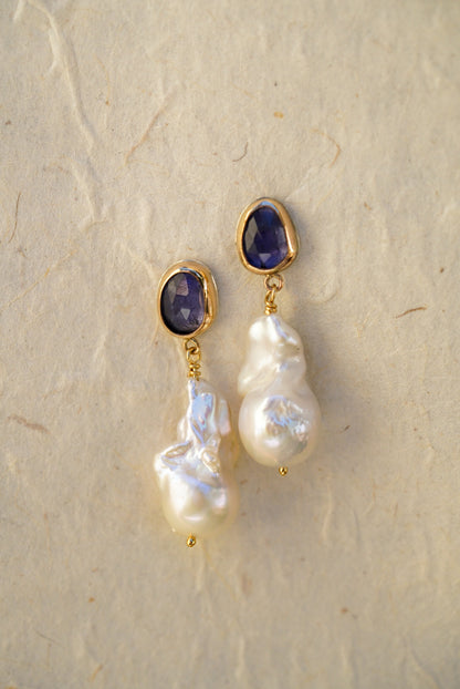 A pair of gold drop earrings with oval tanzanite gemstones at the top and irregular baroque pearl drops below, displayed on a light textured background.